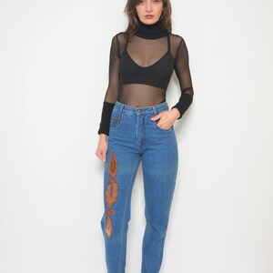 May include: Blue denim jeans with an embroidered design on the left leg. The jeans are paired with a black turtleneck top and a sheer long-sleeved shirt. The outfit is completed with black ankle boots.
