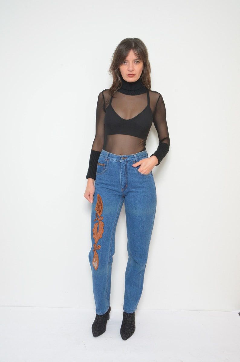 May include: Blue denim jeans with an embroidered design on the left leg. The jeans are paired with a black turtleneck top and a sheer long-sleeved shirt. The outfit is completed with black ankle boots.