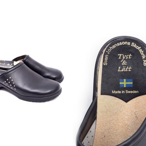 Pode incluir: Um par de tamancos de couro preto com uma bandeira sueca e o texto "Tyst & Lätt" na sola. O texto "Sven Johanssons Skofabrik AB" e "Made in Sweden" também estão impressos na sola.