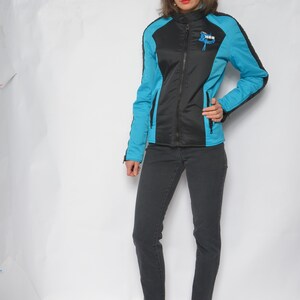 May include: A black and blue jacket with a zipper closure and a logo that says "The Motor" on the left chest. The jacket has a stand-up collar and long sleeves. The model is wearing black jeans and blue shoes.