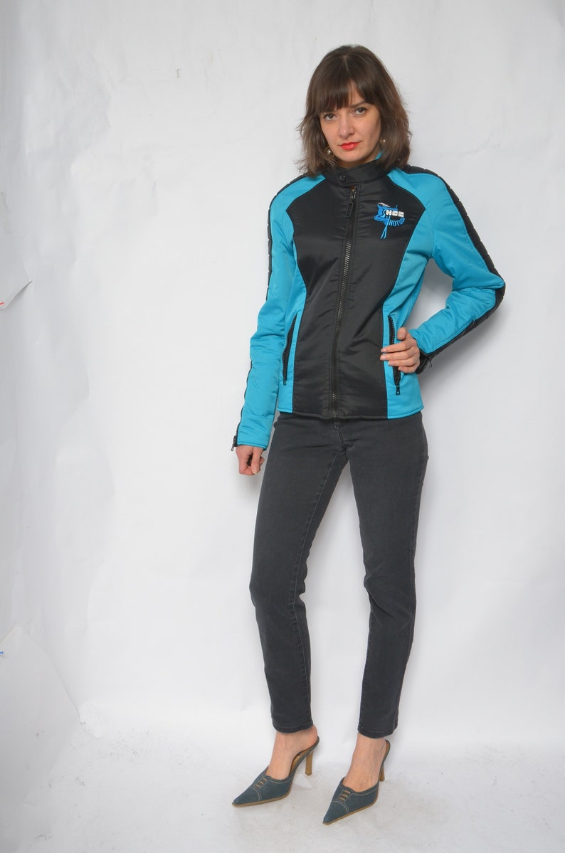 May include: A black and blue jacket with a zipper closure and a logo that says "The Motor" on the left chest. The jacket has a stand-up collar and long sleeves. The model is wearing black jeans and blue shoes.
