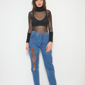 May include: A woman's outfit featuring a black turtleneck mesh top, a black bra, and high-waisted blue jeans. The jeans have an embroidered design on the left leg. The outfit is completed with black wrist warmers and black patterned boots.