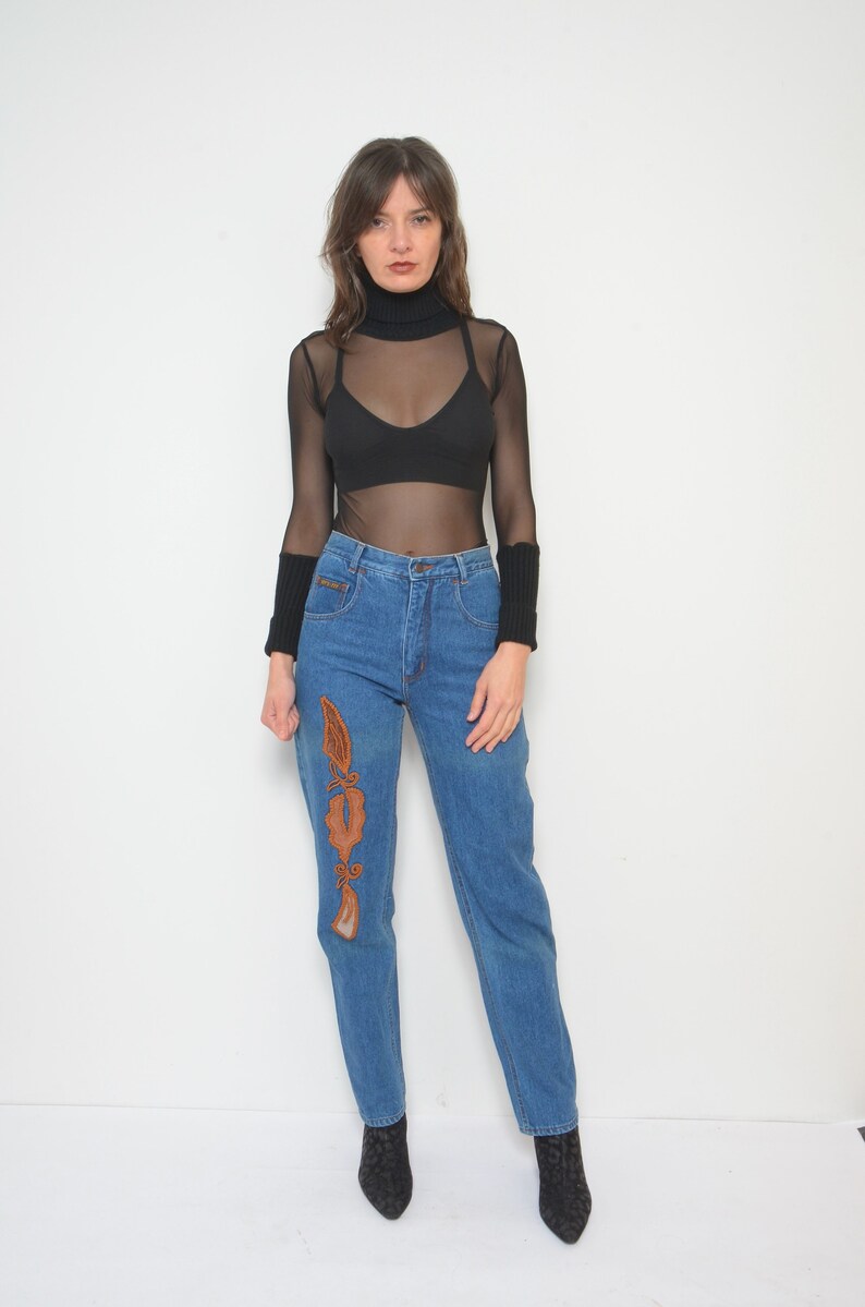 May include: A woman's outfit featuring a black turtleneck mesh top, a black bra, and high-waisted blue jeans. The jeans have an embroidered design on the left leg. The outfit is completed with black wrist warmers and black patterned boots.