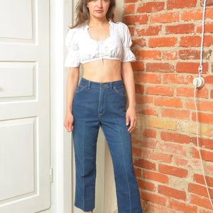 May include: A woman in a white crop top with frilled sleeves and dark blue, high-waisted jeans. She is wearing ankle boots with a snakeskin design. The background features a red brick wall and a white door. The jeans are a classic denim style.