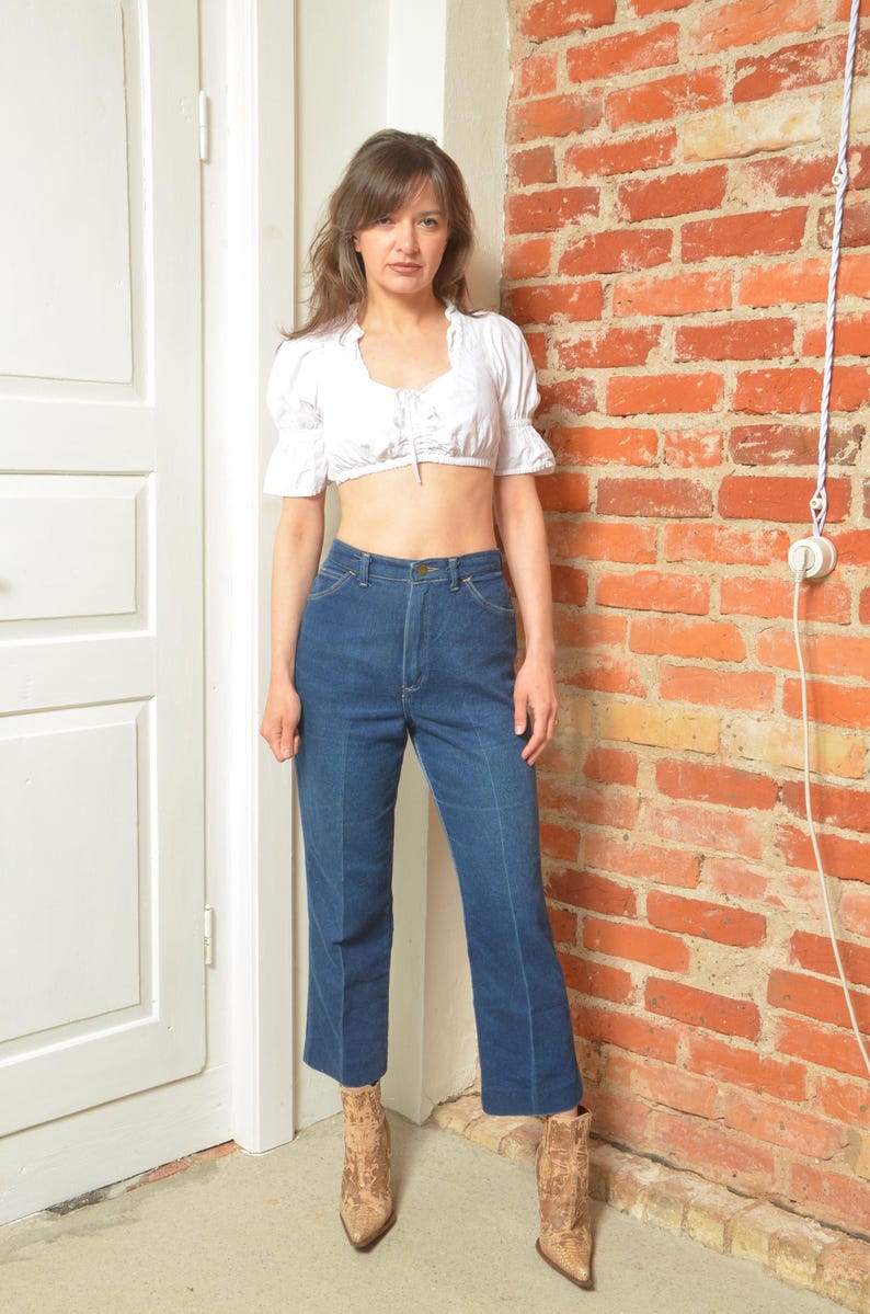 May include: A woman in a white crop top with frilled sleeves and dark blue, high-waisted jeans. She is wearing ankle boots with a snakeskin design. The background features a red brick wall and a white door. The jeans are a classic denim style.