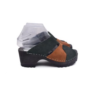May include: Open-toe clogs with a dark wooden sole. The upper is a mix of green and brown suede-like material. The clogs have a block heel and a slip-on design. White stitching details the design. The clogs are a classic style.