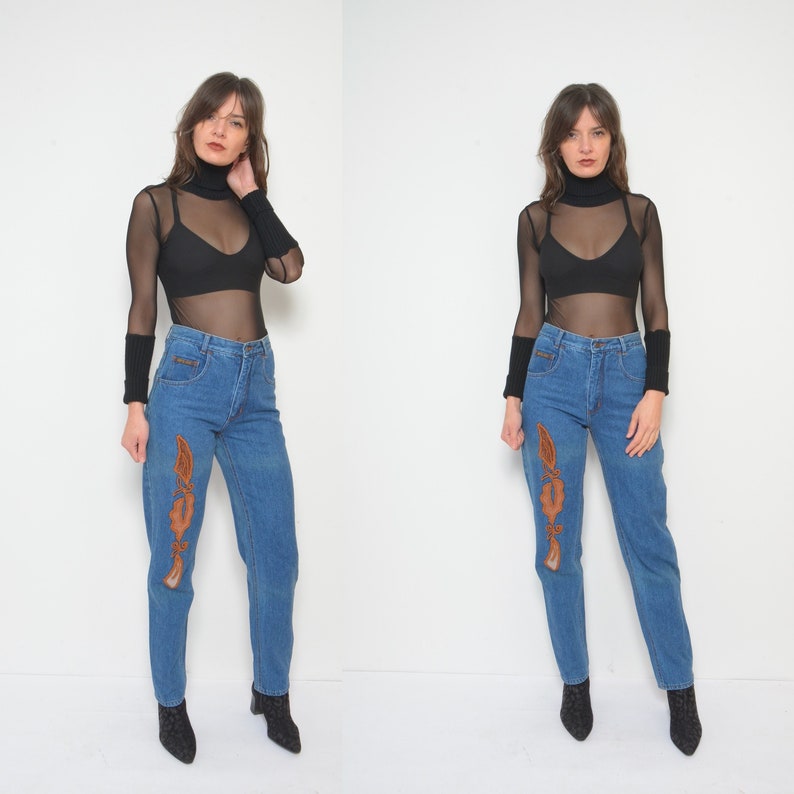 May include: Blue denim jeans with brown decorative stitching on the front. The jeans have a high waist and a straight leg. The model is wearing a black sheer long-sleeve top and black ankle boots.