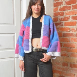 Könnte beinhalten: Eine Häkelstrickjacke mit einem Patchwork-Muster aus blauen, rosafarbenen und violetten Quadraten. Das Kleidungsstück hat lange, weite Ärmel und einen hellgrauen Rippsaum entlang der Vorderöffnung und an den Ärmelbündchen. Die Strickstruktur ist sichtbar.