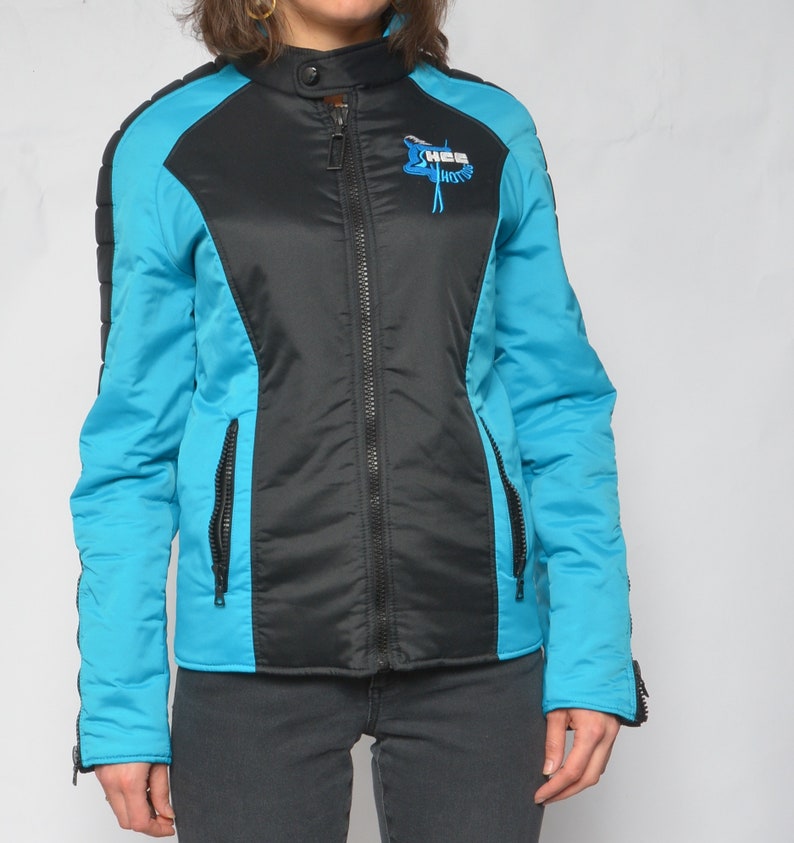 May include: A black and turquoise blue jacket with a zipper closure. The jacket has a logo on the left side that says "X-MAS Motors".