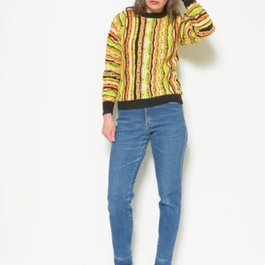 May include: A woman wearing a green, orange, and black striped sweater with a black collar and long sleeves. She is also wearing blue jeans and black heeled ankle boots.