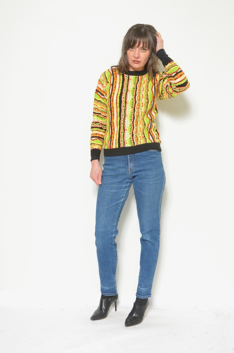 May include: A woman wearing a green, orange, and black striped sweater with a black collar and long sleeves. She is also wearing blue jeans and black heeled ankle boots.