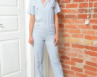 May include: Light blue and white striped jumpsuit with short sleeves and a zip-up front. The jumpsuit features gold-colored zippers on the chest and pockets. The model is wearing orange heels. The background includes a brick wall and a white door.