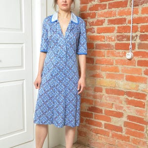 Può includere: Una donna indossa un vestito a motivi blu e bianchi con un colletto bianco e maniche corte. È in piedi davanti a un muro di mattoni e indossa sandali con tacco rosso con decorazioni floreali.