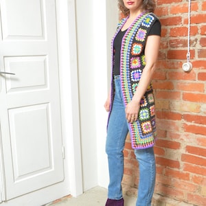 Puede incluir: Un chaleco largo de crochet colorido con un patrón geométrico. El chaleco es sin mangas y tiene cuello en V. La persona que lleva el chaleco está de pie frente a una pared de ladrillo.