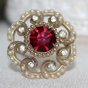 Könnte beinhalten: Vintage-Stil Brosche mit einem zentralen, facettierten, rubinroten Edelstein. Die Brosche ist von kleinen Kunstperlen und klaren Kristallen umgeben, die in einem goldfarbenen Metallrahmen gefasst sind. Das Design ist verziert und floral inspiriert.