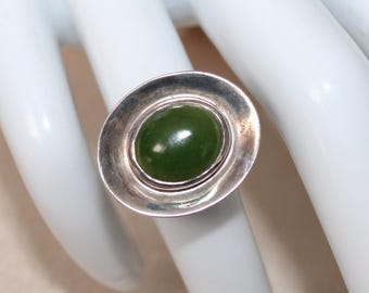 May include: A silver ring featuring an oval, deep green stone. The stone is set in a silver bezel, and the ring is displayed on a white surface. The ring's design is simple and elegant, suitable for everyday wear.