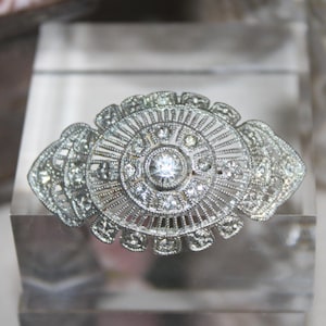 Peut inclure: Broche argentée de style vintage avec une pierre centrale ronde et transparente et des pierres plus petites. La broche présente un motif Art Déco avec un motif en forme d'éventail. La broche est exposée sur un bloc rectangulaire transparent.