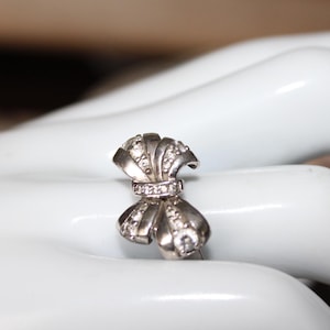 May include: A silver-toned bow-shaped ring with clear gemstones. The ring is set on a white surface, showcasing its intricate design and sparkling details. The bow features a central band of smaller stones.
