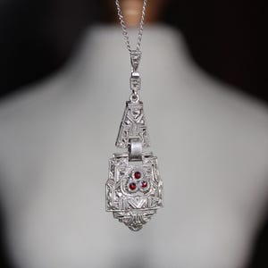 May include: Silver pendant necklace with an ornate, geometric design. The pendant features three red gemstones and hangs from a delicate chain. The necklace is displayed against a blurred background.