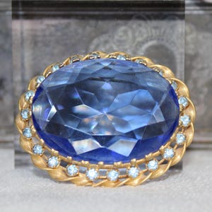 May include: An oval-shaped brooch with a large, faceted blue gemstone at its center. The gemstone is surrounded by a gold-toned frame with a twisted rope design and small, light blue accent stones. The brooch is a vintage style.