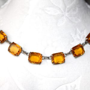 Peut inclure: Un collier vintage orné de pierres carrées de couleur ambre, serties sur une chaîne en métal argenté. Les pierres sont espacées de manière régulière, créant un design symétrique. Le collier est présenté sur une surface blanche.