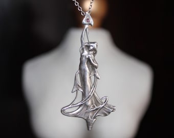 May include: A silver pendant necklace with an Art Nouveau design depicting a woman. The pendant has a flowing, elegant form and a small, pink gemstone. The necklace is shown against a blurred background.
