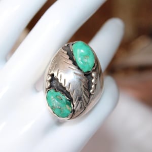 May include: A silver ring with two oval turquoise stones. The ring has a leaf design on the sides and is set on a white surface. The turquoise stones are a bright, vibrant green.
