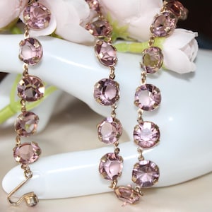 Peut inclure: Un collier délicat orné de pierres précieuses ovales rose clair serties dans une chaîne dorée. Les pierres précieuses sont espacées uniformément et reliées par de petits maillons. Le collier est présenté sur une surface blanche avec des fleurs roses douces en arrière-plan.