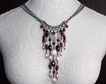 May include: A dark silver-toned necklace with cascading beaded strands. The necklace features a variety of beads, including deep red, clear, and white pearls. The necklace is displayed on a white mannequin.