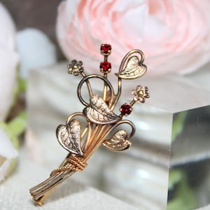 Peut inclure: Broche vintage dorée en forme de bouquet floral. La broche présente des feuilles détaillées, de petites fleurs et des accents de pierres précieuses rouges. Les tiges sont liées avec du fil doré.