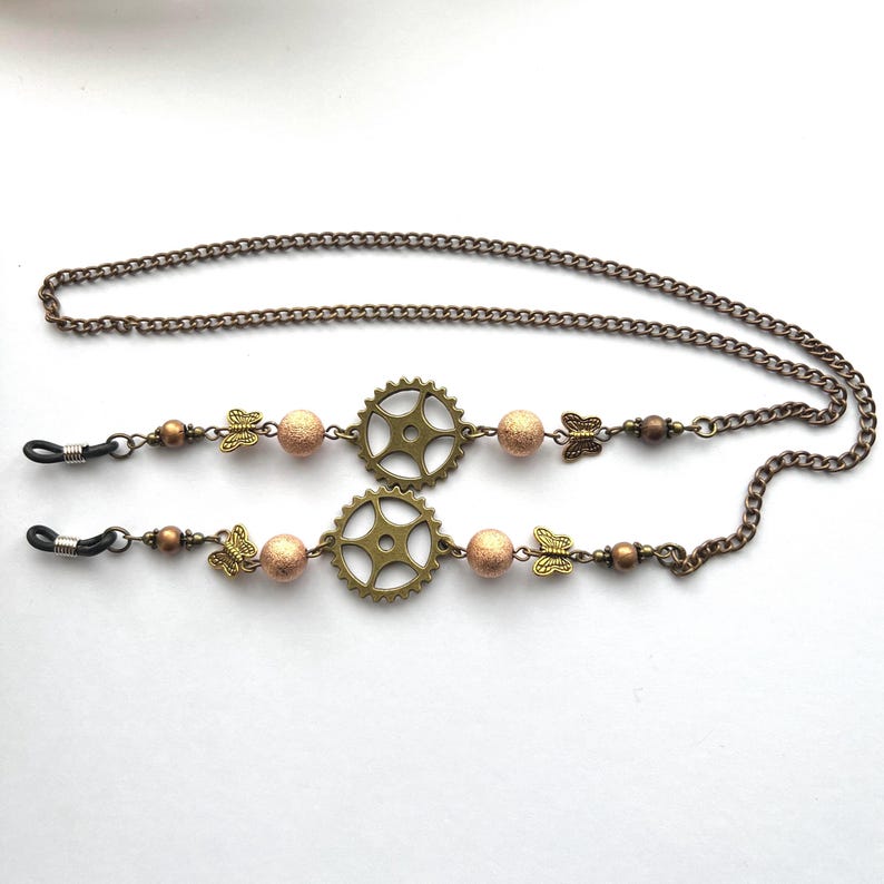 May include: Eyeglass chain with a steampunk aesthetic. The chain is bronze-colored, with a series of small links. The chain features two bronze-colored gear-shaped charms, interspersed with peach-colored beads and butterfly charms.