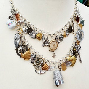 May include: A silver chain necklace with multiple charms, including a small teapot, a compass, a heart, a watch, a key, a thimble, and a feather. The charms are in silver and gold tones.
