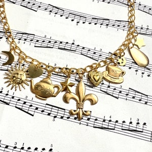 May include: A gold charm bracelet featuring a selection of charms, including a sun, moon, heart, teapot, star, fleur-de-lis, teacup, and a teardrop. The bracelet is displayed on a sheet of music.
