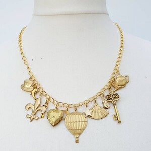 May include: A gold chain necklace with multiple charms, including a heart, a hot air balloon, a teacup, a key, a fleur-de-lis, a star, and a fan.