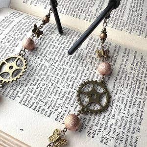 May include: Eyeglass chain with steampunk-style charms. Features a brass gear, butterfly, and textured beads. The chain is shown resting on a book with printed text.