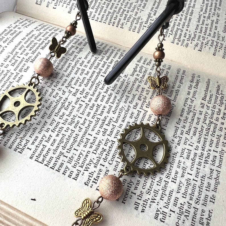 May include: Eyeglass chain with steampunk-style charms. Features a brass gear, butterfly, and textured beads. The chain is shown resting on a book with printed text.