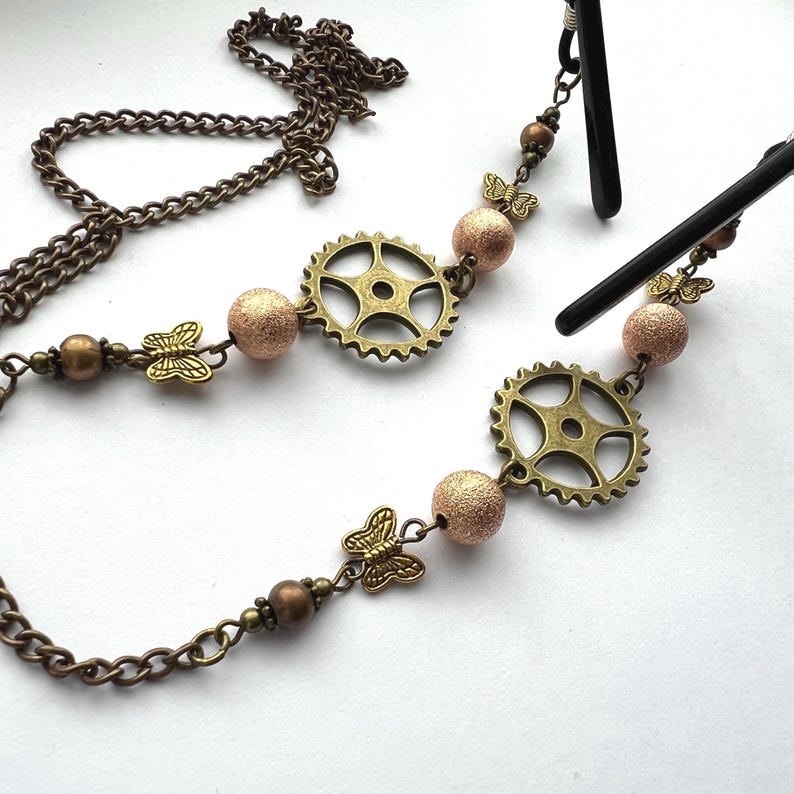 May include: A steampunk-style eyeglass chain with a bronze chain, gear-shaped charms, butterfly accents, and textured pink beads. The chain is designed to hold eyeglasses and has a vintage aesthetic.