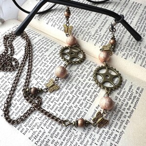 May include: Antique bronze necklace with a chain, adorned with butterfly charms, gear-shaped pendants, and textured pink beads. The necklace is displayed on an open book.