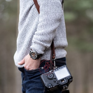 May include: A person wearing a grey sweater and blue jeans with a brown leather camera strap. A black camera with a large lens is attached to the strap.