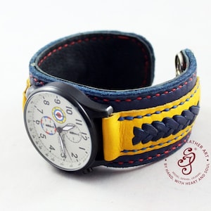 May include: A watch with a white face and black hands, featuring a red, white, and blue design. The watch is attached to a yellow and blue leather cuff bracelet with red stitching. The bracelet has a braided design and the text "Leather Art. By Hand, With Heart and Soul." is printed on the side.
