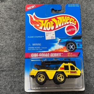 May include: A yellow toy fire truck with a black ladder and a black and red "Fire Stopper" logo. The truck is part of the "Fire Squad Series" and is number 3 of the series. The packaging is blue and red with the Hot Wheels logo.