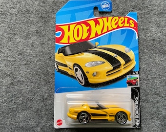 Puede incluir: Un coche de juguete amarillo de fundición a presión Hot Wheels modelo Dodge Viper Roadster. El coche tiene rayas de carreras negras y un interior negro. El coche está sobre un fondo azul con el logotipo de Hot Wheels y las palabras "HW Roadsters".