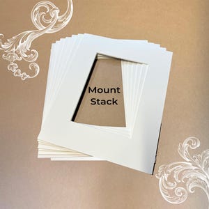 May include: A stack of white rectangular picture frame mounts with a central rectangular aperture. The text Mount Stack is printed in black in the centre of the opening. The mounts are arranged in a layered pile on a tan background.