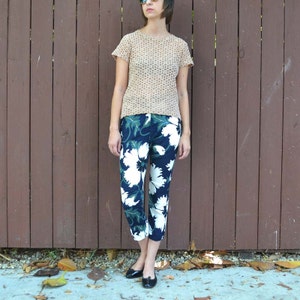 May include: A woman wearing a beige crocheted top and blue and white floral cropped pants. She is standing in front of a brown wooden fence.