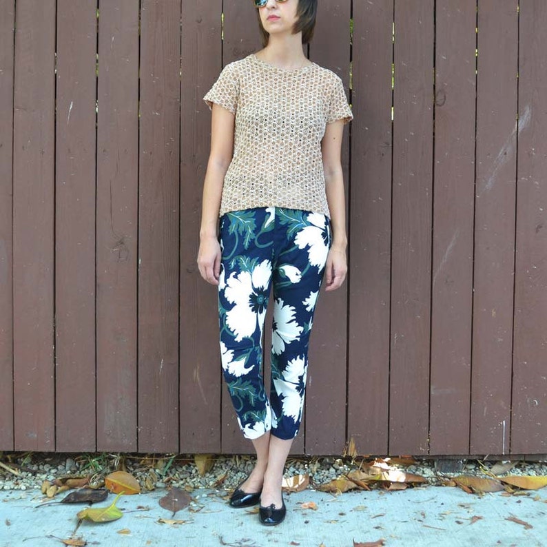 May include: A woman wearing a beige crocheted top and blue and white floral cropped pants. She is standing in front of a brown wooden fence.