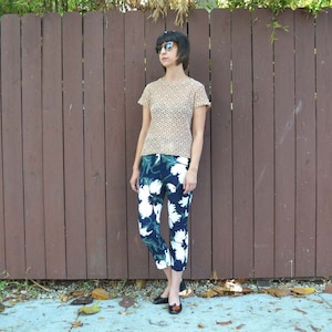 May include: A woman wearing a beige crochet top with a floral pattern and dark blue cropped pants. She is wearing sunglasses and has short dark hair.