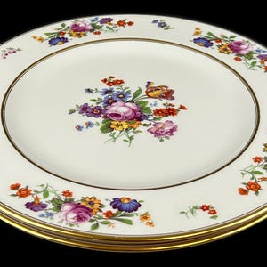 May include: A stack of fine china plates with a cream-colored surface. Each plate features a gold rim and a floral design with pink, purple, orange, and blue flowers. The plates are stacked on a black background.