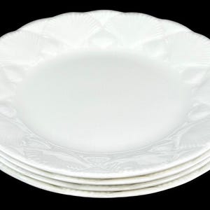 May include: A stack of three white ceramic plates with a smooth, circular center and a decorative scalloped rim featuring a raised, embossed shell pattern that extends around the entire edge of each plate, creating a textured border.