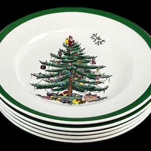 May include: A stack of white ceramic plates with a dark green rim. The center of each plate features a detailed illustration of a decorated Christmas tree with colorful ornaments, a Santa Claus figure at the top, and wrapped gifts at the base.
