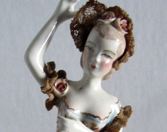 May include: A porcelain figurine of a woman wearing a brown lace dress and a floral headpiece. She is holding her arm up and has a painted floral design on her dress.
